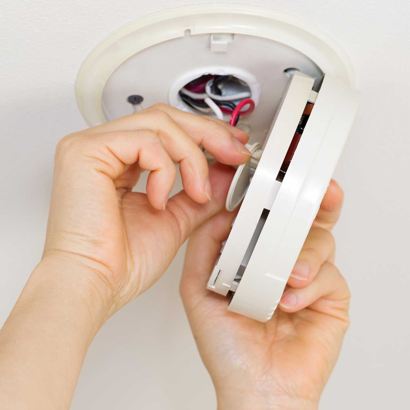 Doing Maintenance on Home Smoke Dectector. Horizontal photo of female hands taking home smoke detector apart with white ceiling background